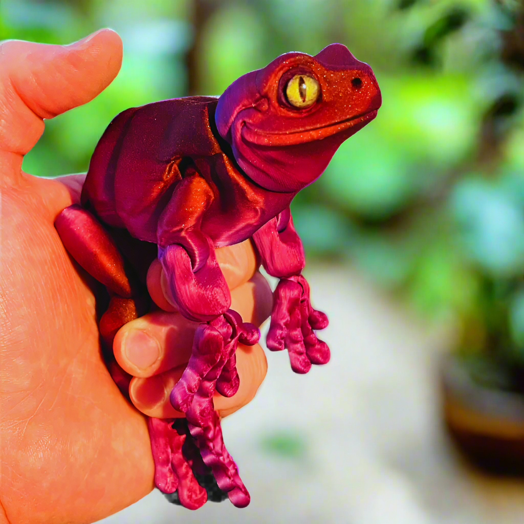 A 3D printed red and purple tree frog held in a hand to display its size, featuring large eyes and a smooth, detailed surface.