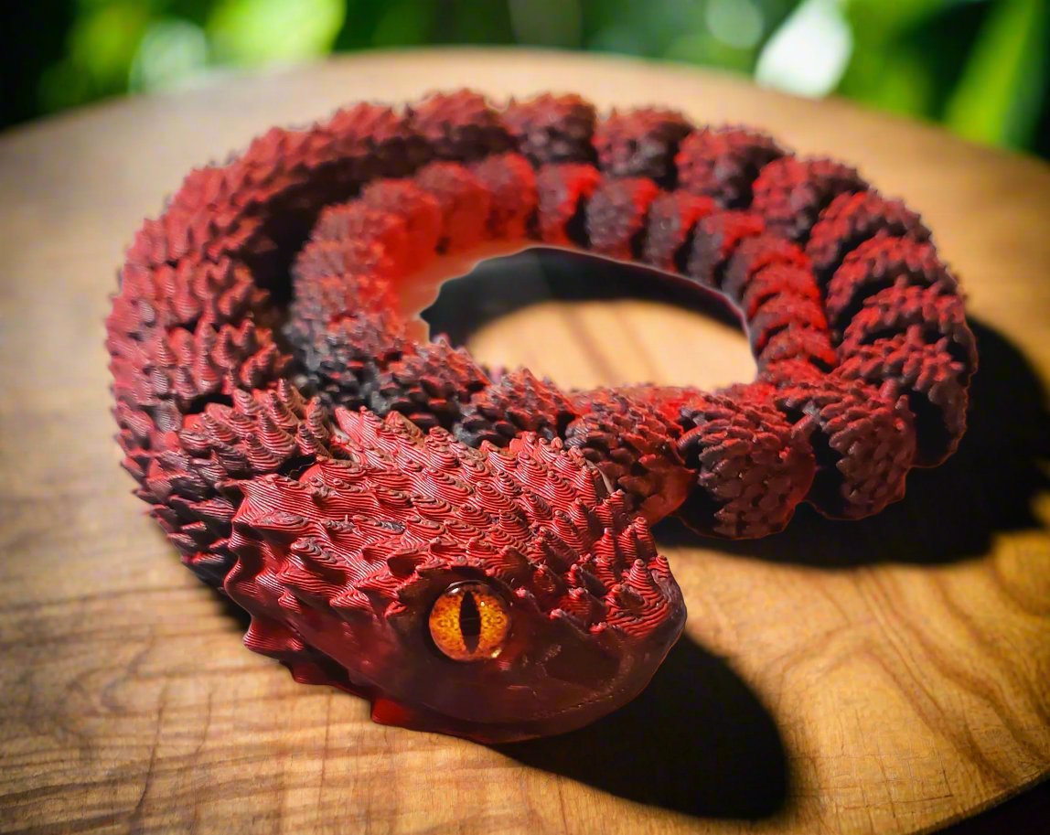 A red and black coiled, intricately textured snake figure with a bright amber eye, displayed on a light surface.